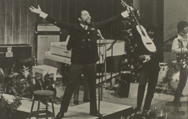 Gilberto_Gil_nos_anos_1960 em um palco com os braços abertos segurando um violão